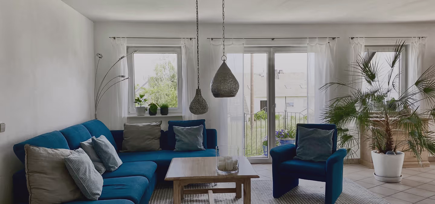 Modernes Wohnzimmer mit blauem Sofa und Sessel, Holztisch, großen Fenstern mit weißen Vorhängen und Zimmerpflanzen.